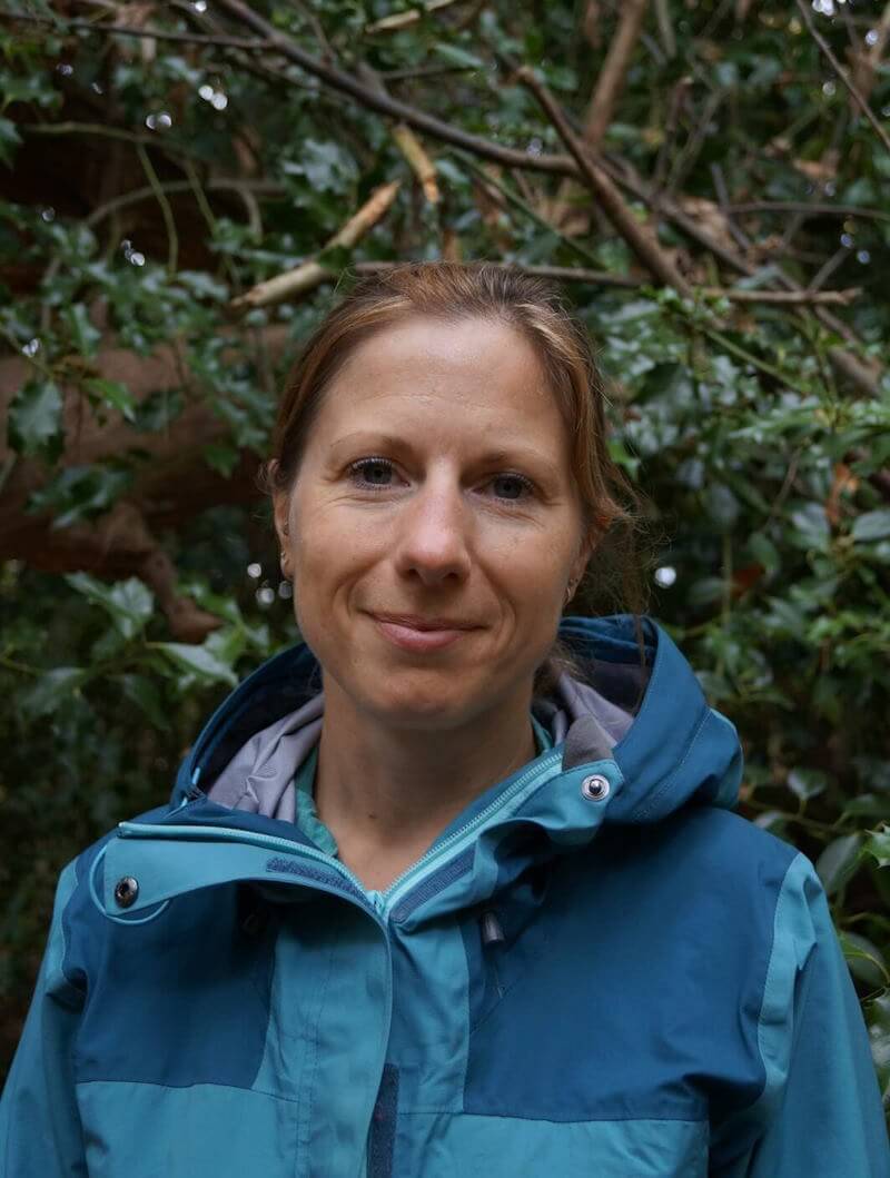 A portrait photograph of Liz Cramp therapist in a forest, wearing a blue anorak